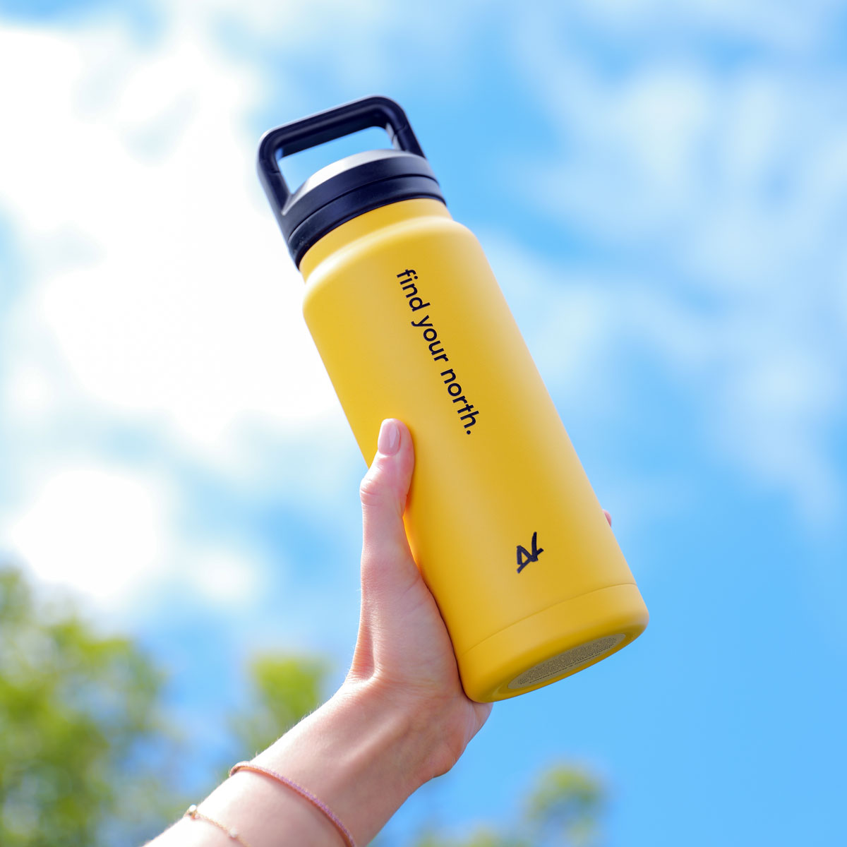 A hand holding a bright yellow stainless steel water bottle with a black loop handle and black printed text reading 'find your north.' against a bright blue sky background.